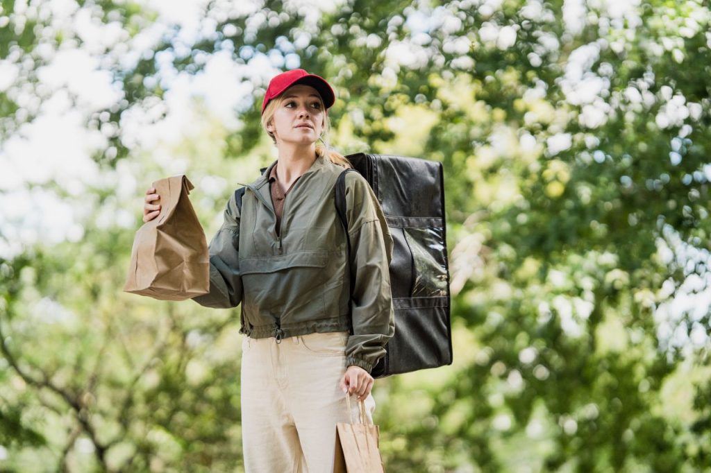 A woman in a red cap delivers meals in a park, holding paper bags.